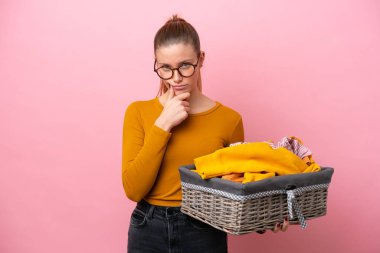Young caucasian woman holding a clothes basket isolated on pink background thinking