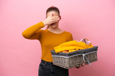 Young caucasian woman holding a clothes basket isolated on pink background covering eyes by hands. Do not want to see something