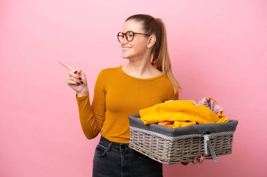 Young caucasian woman holding a clothes basket isolated on pink background pointing finger to the side and presenting a product
