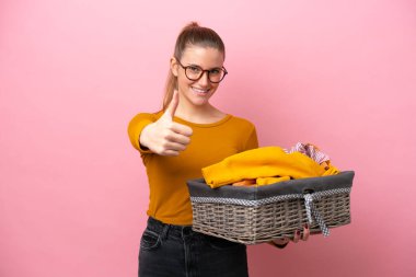 Young caucasian woman holding a clothes basket isolated on pink background with thumbs up because something good has happened
