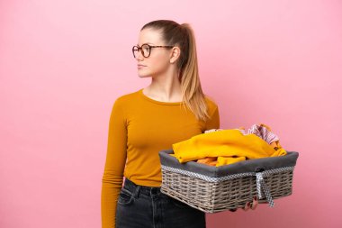 Young caucasian woman holding a clothes basket isolated on pink background looking to the side