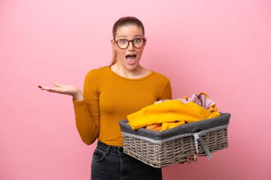 Young caucasian woman holding a clothes basket isolated on pink background with shocked facial expression