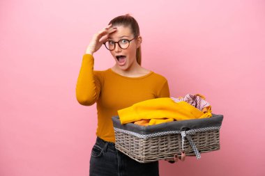 Young caucasian woman holding a clothes basket isolated on pink background doing surprise gesture while looking to the side