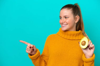 Young caucasian woman holding an avocado isolated on blue background pointing to the side to present a product