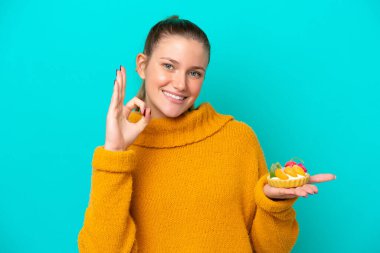 Young caucasian woman holding a tartlet isolated on blue background showing ok sign with fingers