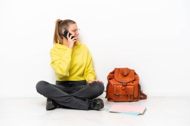 Beyaz arka planda bir katta oturan beyaz tenli genç bir kadın cep telefonuyla sohbet ediyor.