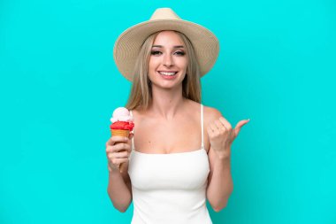 Mavi arka planda izole edilmiş dondurma tutan mayolu sarışın bir kadın bir ürünü sunmak için yan tarafı işaret ediyor.