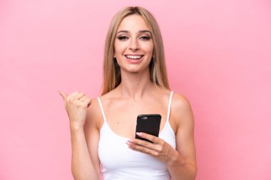 Pretty blonde woman isolated on pink background using mobile phone and pointing to the lateral