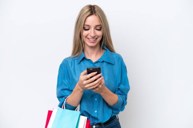 Pretty blonde woman isolated on white background holding shopping bags and writing a message with her cell phone to a friend