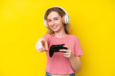 Blonde English young girl playing with a video game controller isolated on yellow background pointing front with happy expression