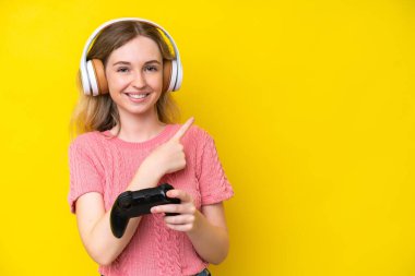 Blonde English young girl playing with a video game controller isolated on yellow background pointing to the side to present a product