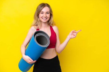 Young sport caucasian woman going to yoga classes while holding a mat isolated on yellow background pointing finger to the side