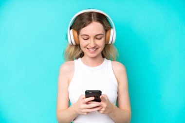 Blonde English young girl isolated on blue background listening music and looking to mobile