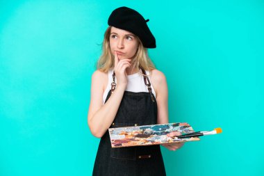 Young artist caucasian woman holding a palette isolated on blue background having doubts while looking up