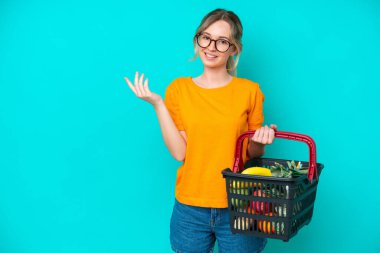 Blonde English young girl holding a shopping basket full of food isolated on blue background extending hands to the side for inviting to come