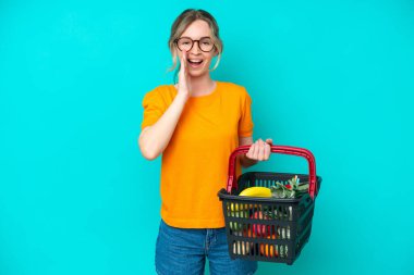 Blonde English young girl holding a shopping basket full of food isolated on blue background shouting with mouth wide open