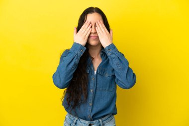 Sarı arka planda izole edilmiş genç, beyaz bir kadın elleriyle gözlerini kapatıyor.