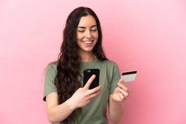 Pembe arka planda izole edilmiş genç beyaz bir kadın kredi kartıyla cep telefonuyla alışveriş yapıyor.
