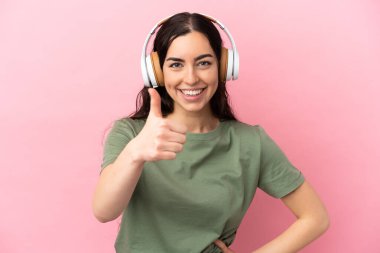 Genç beyaz kadın pembe arka planda izole edilmiş müzik dinliyor ve başparmağını kaldırıyor.