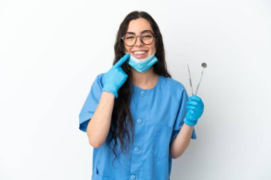 Beyaz arka planda izole edilmiş aletleri tutan genç dişçi kadın baş parmak işareti yapıyor.