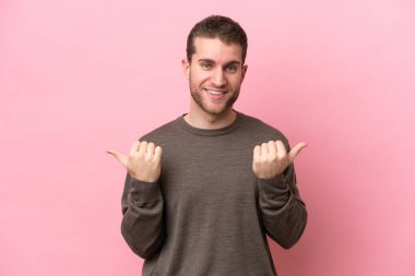 Pembe arka planda izole edilmiş genç beyaz bir adam. Baş parmakları havada ve gülümsüyor.