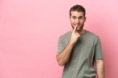 Pembe arka planda izole edilmiş genç beyaz adam sessiz bir jest işareti gösteriyor. Parmağını ağzına sokuyor.