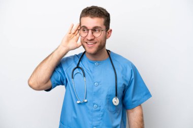 Young surgeon doctor caucasian man isolated on white background listening to something by putting hand on the ear