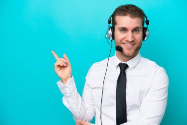 Telemarketer caucasian man working with a headset isolated on blue background pointing finger to the side