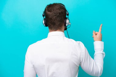 Telemarketer caucasian man working with a headset isolated on blue background pointing back with the index finger