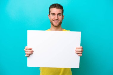 Young caucasian man isolated on blue background holding an empty placard with happy expression