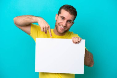 Young caucasian man isolated on blue background holding an empty placard with happy expression and pointing it