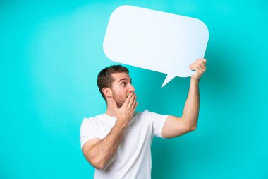 Kafkasyalı genç adam mavi arka planda izole edilmiş. Elinde şaşırmış bir ifadeyle boş bir konuşma balonu tutuyor.