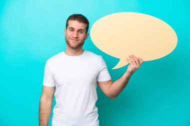 Mavi arka planda izole genç beyaz adam boş bir konuşma balonu tutuyor.