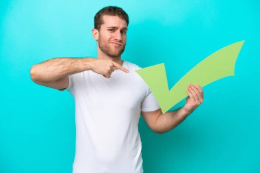 Kafkasyalı genç adam mavi arka planda izole edilmiş bir şekilde elinde bir kontrol simgesi tutuyor ve ona doğrultuyor.