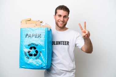 Beyaz arka planda izole edilmiş geri dönüşüm için kağıt dolu bir çanta taşıyan beyaz bir adam gülümsüyor ve zafer işareti gösteriyor.