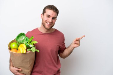 Young caucasian man holding a grocery shopping bag isolated on white background pointing finger to the side