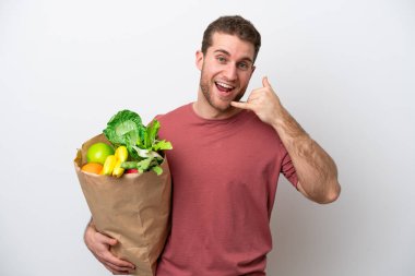 Young caucasian man holding a grocery shopping bag isolated on white background making phone gesture. Call me back sign