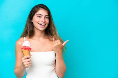 Young woman in swimsuit holding an ice cream isolated on blue background pointing to the side to present a product