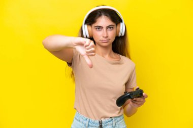 Young caucasian woman playing with a video game controller isolated on yellow background showing thumb down with negative expression