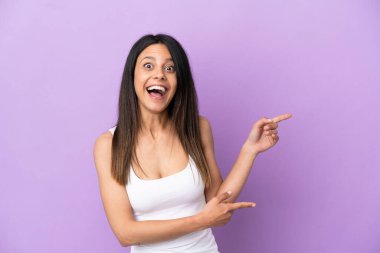 Mor arka planda izole edilmiş genç beyaz kadın şaşırmış ve işaret ediyor.