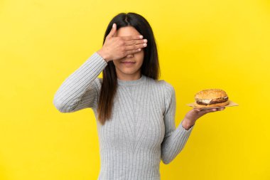 Young caucasian woman holding a burger isolated on yellow background covering eyes by hands. Do not want to see something