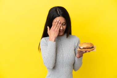 Young caucasian woman holding a burger isolated on yellow background with tired and sick expression