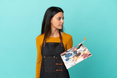 Young artist caucasian woman holding a palette isolated on blue background looking to the side