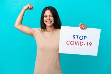 Young woman over isolated background holding a placard with text Stop Covid 19 and doing strong gesture