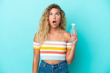 Young blonde woman with a bottle of water isolated on blue background looking up and with surprised expression