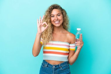 Young blonde woman with a bottle of water isolated on blue background showing ok sign with fingers