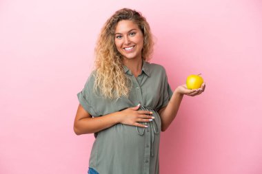 Kıvırcık saçlı kız pembe arka planda izole edilmiş hamile ve elinde elma var.