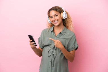 Kıvırcık saçlı kız pembe arka planda izole edilmiş hamile ve cep telefonuyla müzik dinliyor.