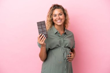 Kıvırcık saçlı kız pembe arka planda izole edilmiş hamile ve elinde çikolata var.