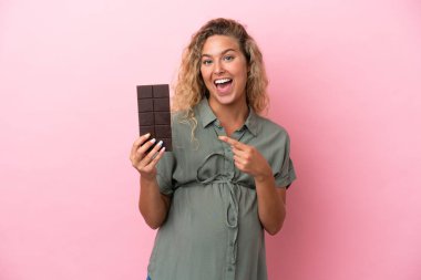 Kıvırcık saçlı kız pembe arka planda izole edilmiş hamile ve elinde çikolata var.